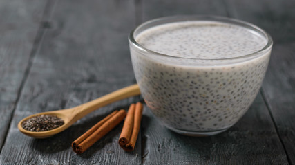 Black Chia seed pudding in a glass bowl, wooden spoon and cinnamon sticks on a black wooden table.