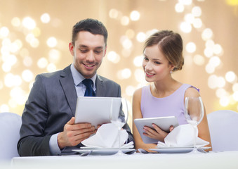 dating, luxury and people concept - happy couple with menus on tablet pc computers at restaurant over festive lights on beige background