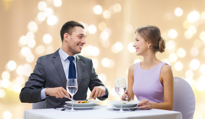 people and leisure concept - smiling couple eating appetizers at restaurant over festive lights on beige background