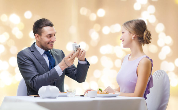 couple, technology and leisure concept - smiling man taking picture of wife or girlfriend by digital camera at restaurant over festive lights on beige background