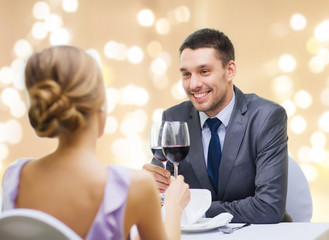 dating, celebration and valentines day concept - smiling young couple clinking glasses of non-alcoholic red wine and looking at each other at restaurant over festive lights on beige background