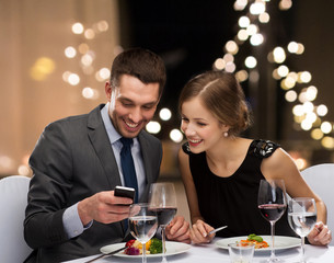 technology, leisure and people concept - happy couple with smartphone eating at restaurant over festive lights on background