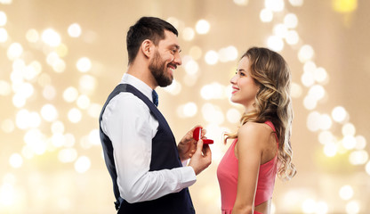 love, couple, proposal and people concept - happy man giving diamond engagement ring in little red box to woman over festive lights background