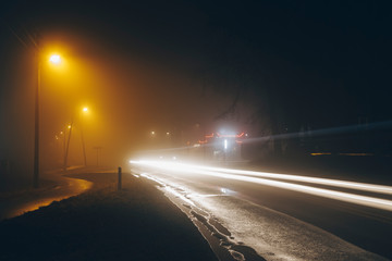 White car light trails. Street light is yellow or orange. Foggy and rainy weather. There are puddles on the pavement. Empty space. Night. Spring or autumn. 