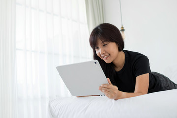 Smiling Asian woman using tablet for video conversation while relaxing on bed in home. Concept of young business people working at home. Copy space.