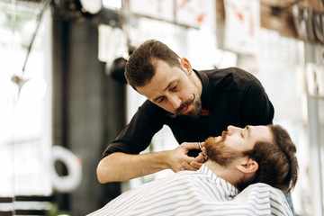 Fashion barber with mustache dressed in a black shirt with a red bow tie tidies up men's beard  and scissors it in the barbershop
