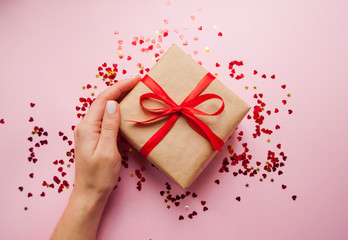 Gift box wrapped in brown colored craft paper and tied with red bow on pink background with red confetti. Lady's hand holding a giftbox.