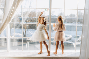 Two little sisters dressed in the beautiful dresses stand on the windowsill next to the mirror