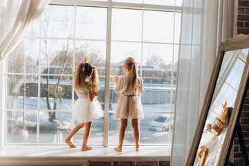 Two little sisters dressed in the beautiful dresses stand on the windowsill next to the mirror