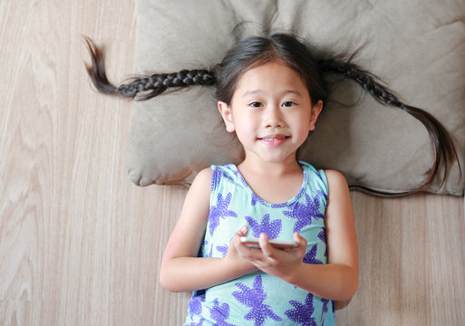 Happy Asian child girl playing smartphone lying on wooden floor. Top view.