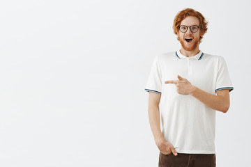 Indoor shot o fimpressed speechless funny ginger man with long beard gasping and smiling with excitement pointing left raising shoulders while seeing interesting and amazing thing over gray wall