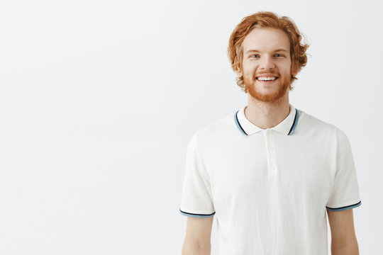 Waist-up shot of carefree ordinary redhead bearded man with messy stylish hairstyle in white polo shirt smiling with casual friendly and happy smile enjoying great summer day over gray background