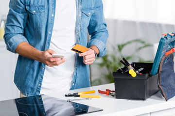 cropped view of man holding smartphone and credit card near tool box