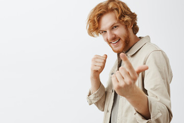 Close-up shot of confident playful and happy reckless redhead man with beard and wavy hair inviting with index finger to fight raising fist wanting to start argument over gray wall