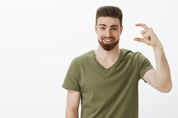 Attractive upbeat bearded sportsman in olive t-shirt shape small or tiny object telling about little effort till goal, smiling broadly with assured and confident expression at camera over white wall