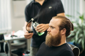 Fototapeta premium The hands of young barber making haircut to attractive bearded man in barbershop