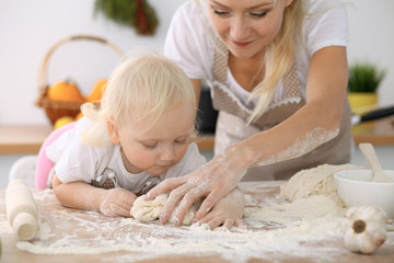 Little girl and her blonde mom in beige aprons  playing and laughing while kneading the dough in kitchen. Homemade pastry for bread, pizza or bake cookies. Family fun and cooking concept