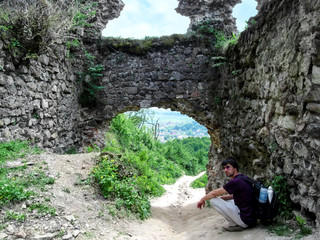Tourist with a backpack and sunglasses squatted near the remnants of the Khust Castle Gate. A young man rests near the stone entrance to the medieval ruined fortress on a clear sunny spring-summer day