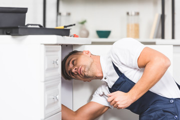 adult repairman holding spanner while checking pipes at kitchen