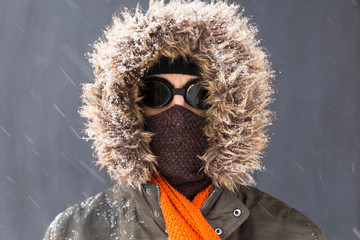 Portrait of a single male winter adventurer wearing a warm green coat with fur hood, a blue ski cap, an orange scarf and black retro style goggles