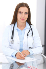 Doctor woman at work. Portrait of female physician filling up medical form while sitting at the glass desk at clinic or hospital. Medicine and healthcare concept