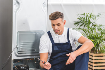Fototapeta premium confused adult repairman looking at screwdriver while repairing refrigerator