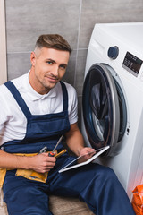 Obraz premium adult repairman holding screwdriver and using digital tablet while repairing washing machine in bathroom and looking at camera