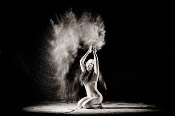 The girl with the flour on the body stretches the arms up with thrown flour on black background black and white image