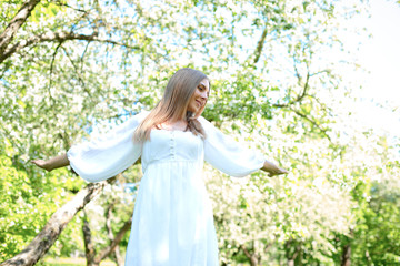 Fototapeta na wymiar Happy blonde woman on a background of blooming spring garden with her hands raised to the sky
