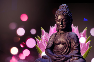 Buddha statue sitting in meditation pose against deep dark background and pink lotus flower behind.