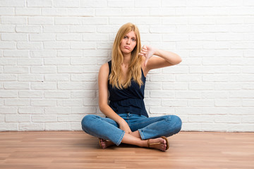 Fototapeta na wymiar Blonde girl sitting on the floor showing thumb down sign with negative expression on white brick wall background