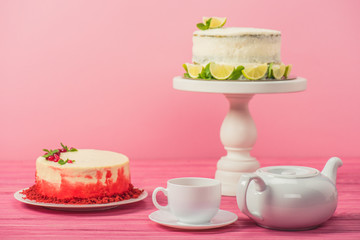 cakes decorated with currants, mint leaves and lime slices near cup and tea pot isolated on pink