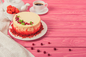 close up of cake decorated with currants and mint leaves near tea pot and cup of strawberries on pink surface
