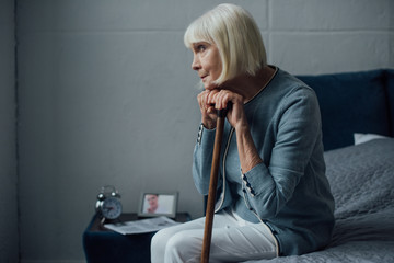lonely senior woman with hands on walking stick sitting on bed at home