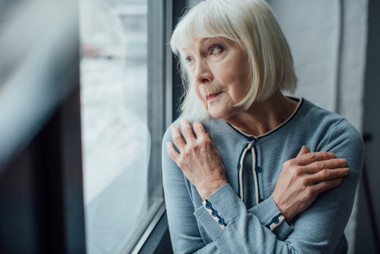 pensive senior woman with hands crossed looking through window at home