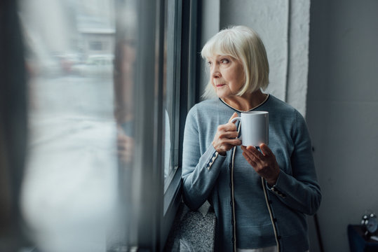 Pensive senior woman holding cup of coffee and looking through window at home