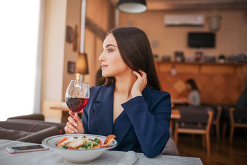 Beautiful young woman sit at table and look to left. She hold glass of red wine and touch her hair. Salad bown stand in front of her. Phone lying besides.