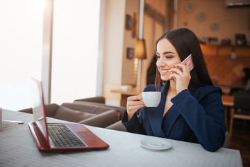 Cheerful young woman sit at table and work. She smiles. Customer talk on phone and look at laptop's screen. Young woman drink coffee from cup.