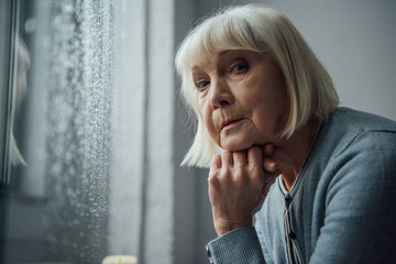 thoughtful senior woman with grey hair propping chin with hand and looking at camera at home