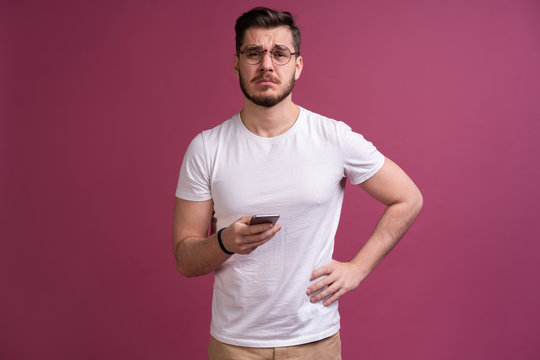Always in touch. Smiling young man holding smart phone and looking at it. Portrait of a happy man using mobile phone isolated over pink background