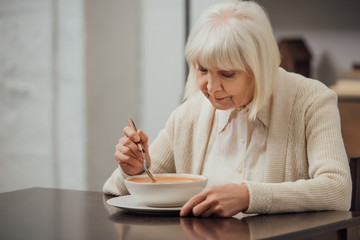 senior woman with grey hair sitting at table and eating cream soup at home