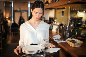 Serious young wautress look down. She hold plates on tray. Waitress stand at bar.