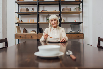 selective focus of upset senior woman eating at table with bowl and spoon on foreground