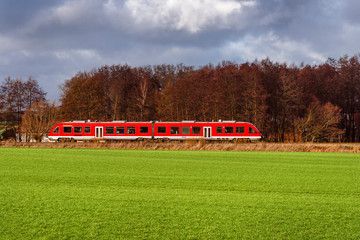 Red modern commuter train with wide landscape, green field, trees and dramatic cloudy sky - concept travel tourism local regional railway traffic transport locomotive departure arrival journey