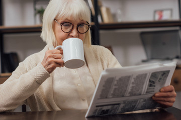 senior woman in glasses sitting at table and reading newspaper while drinking coffee at home