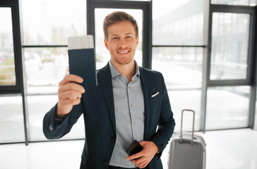 Happy young buisnessman stand in airport hall and show passport with ticket. He looks on camera and smile. Guy has phone in left hand. His suitcase stand behind him.