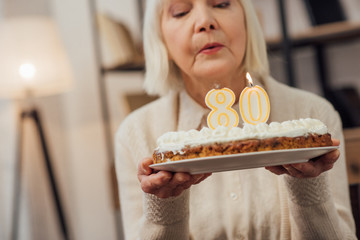 senior woman blowing out candles on cake with number 80 on top while celebrating birthday at home