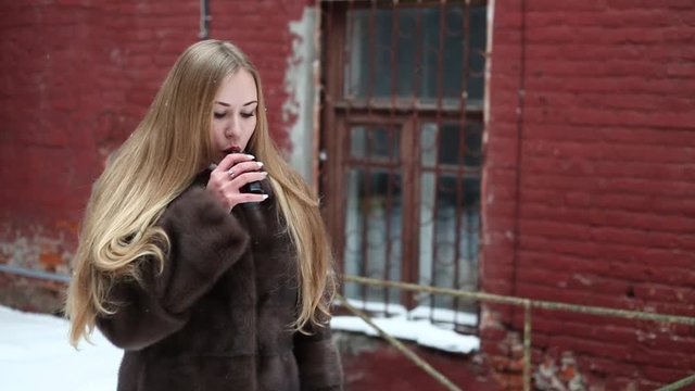 Vape lady. Young pretty girl blonde in a fur coat smokes an electronic cigarette and lets off puffs of steam in a vintage yard outdoors in the winter.