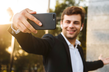 Photo of office worker in suit holding smartphone for selfie, while standing outdoor against building