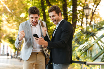 Image of happy businessmen in suits using smartphone while walking outdoor through green park with takeaway coffee, during sunny day